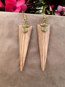 Her Best Foot Forward’s Rosemiti Hand-Carved Rosewood Earrings feature long, triangular shapes with brass accents. This bold statement jewelry from East Africa is artfully shown on a brown surface with pink flowers in the background.