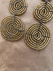 The Kiungo Conjoined Circle Earrings by Her Best Foot Forward feature spiral gold beads and black wire in a handmade Maasai statement design, showcasing unique East African jewelry on a brown textured surface.