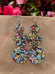 The Kiungo Conjoined Circle Earrings by Her Best Foot Forward are handmade Maasai statement jewelry, featuring colorful beaded rings, displayed on brown paper with vibrant pink flowers and green leaves in the background.