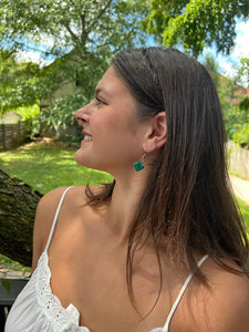 A woman with long brown hair in a white spaghetti strap top smiles outdoors, wearing Her Best Foot Forwards Kijani Malachite Square Earrings—handmade green stone jewelry from East Africa—with trees in the background.