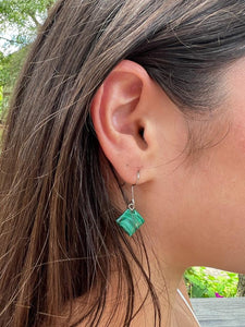 A woman with long brown hair wears Her Best Foot Forward’s Kijani Malachite Square Earrings, handmade green stone jewelry from East Africa, the silver and green design popping against blurred outdoor greenery.