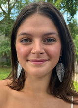 Load image into Gallery viewer, A young woman with long brown hair smiles outdoors, wearing Chuma Etched Metal Earrings by Her Best Foot Forward—lightweight handmade statement jewelry from East Africa. Green trees and grass create a soft background.