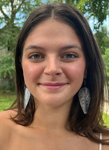 A young woman with long brown hair smiles outdoors, wearing Chuma Etched Metal Earrings by Her Best Foot Forward—lightweight handmade statement jewelry from East Africa. Green trees and grass create a soft background.