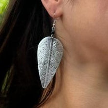 Load image into Gallery viewer, A person with long dark hair wears a Chuma Etched Metal Earring by Her Best Foot Forward—a large, lightweight, handmade statement piece from East Africa with a silver, leaf-shaped textured design hanging from a hook.