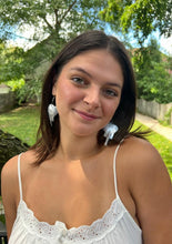 Load image into Gallery viewer, A young woman with shoulder-length brown hair, wearing a white spaghetti-strap top and Her Best Foot Forward’s Chuma Etched Metal Earrings—lightweight, handmade statement jewelry from East Africa—smiles softly in a sunlit green garden.