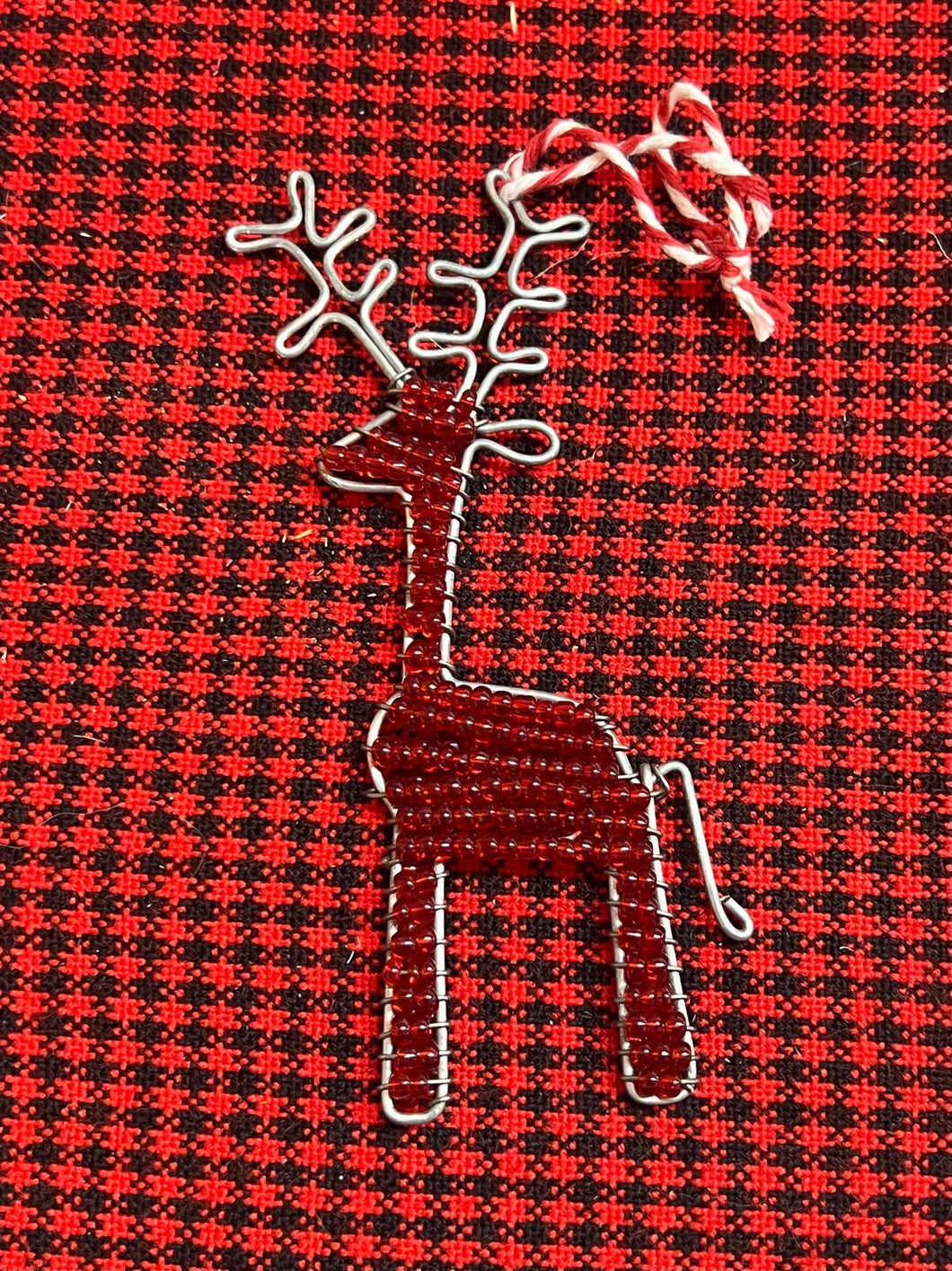 The Reindeer Friends ornament by Her Best Foot Forward is a handcrafted, beaded holiday decoration from Tanzania featuring detailed antlers and a hanging loop, displayed on red and black checkered fabric.