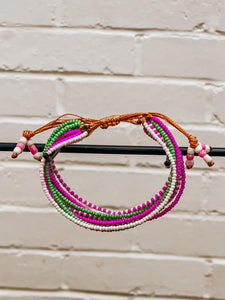 The Shanga Slide Beaded Bracelet by Her Best Foot Forward, an easy slip-on Maasai jewelry piece from East Africa, features pink, green, and white beads on multistrand brown adjustable cords, displayed hanging on a rod against a white brick wall.