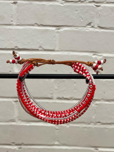 The Shanga Slide Beaded Bracelet by Her Best Foot Forward features red and white beads on a brown adjustable cord, inspired by Maasai jewelry from East Africa, displayed on a horizontal black rod with a white brick wall background.