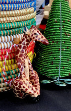 Load image into Gallery viewer, A Wooden Holiday Friends ornament from Her Best Foot Forward—a handcrafted giraffe from Tanzania—sits near a green beaded Christmas tree, reflecting African artistry, with vibrant woven baskets behind.