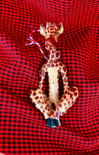 Load image into Gallery viewer, The Wooden Holiday Friends ornament by Her Best Foot Forward is a handcrafted giraffe figurine from Tanzania, featuring a patterned body and sitting cross-legged on vibrant red and black checked fabric.
