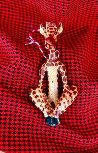 The Wooden Holiday Friends ornament by Her Best Foot Forward is a handcrafted giraffe figurine from Tanzania, featuring a patterned body and sitting cross-legged on vibrant red and black checked fabric.