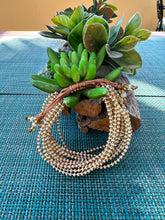 Load image into Gallery viewer, The Shanga Slide Beaded Bracelet by Her Best Foot Forward, inspired by Maasai jewelry, features white and gold beads and is showcased on a textured rock with succulent plants atop a blue woven surface.
