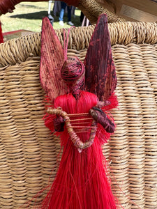 The Angels From On High ornament by Her Best Foot Forward is a handcrafted Christmas angel from Tanzania, made with red and maroon fibers, large dyed wings, and a twisted cord, displayed on a woven basket background.