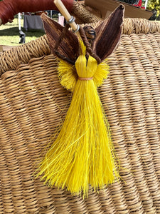 The Angels From On High ornament by Her Best Foot Forward features a handmade yellow banana fiber angel with brown leaf wings on a woven basket, crafted by Tanzanian women artisans, shown outdoors on green grass.
