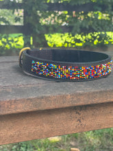 Load image into Gallery viewer, The Buddy Beaded Dog Collar by Her Best Foot Forward, a classic handmade Maasai pet accessory from East Africa, features colorful beadwork on dark leather and rests outdoors on weathered wood with blurred green foliage in the background.