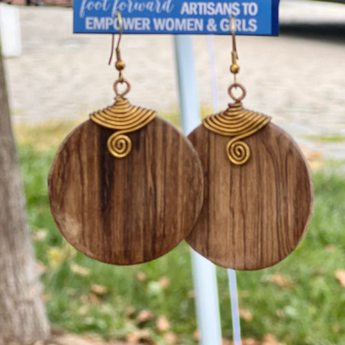 The Rosemiti Hand-Carved Rosewood Earrings by Her Best Foot Forward feature large, round wooden drops with golden brass spiral accents. In the background, a sign reads ARTISANS TO EMPOWER WOMEN & GIRLS.