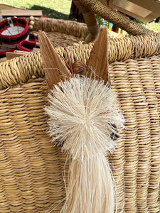 A close-up of the Angels From On High ornament by Her Best Foot Forward, handcrafted in Tanzania from banana fiber with wooden wings, set against a wicker basket. Other baskets woven by Tanzanian women artisans are blurred in the background.