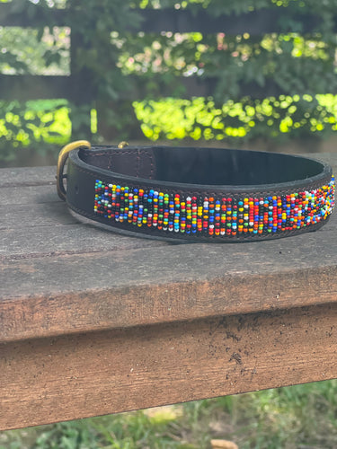 The Buddy Beaded Dog Collar by Her Best Foot Forward, a classic handmade Maasai pet accessory from East Africa, features colorful beadwork on dark leather and rests outdoors on weathered wood with blurred green foliage in the background.