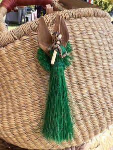 The Angels From On High handcrafted fiber Christmas ornament by Her Best Foot Forward is displayed on a large woven straw basket, accented with two wooden leaves and green raffia. The background shows a blurred outdoor setting.