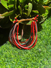 Load image into Gallery viewer, The Shanga Slide Beaded Bracelet by Her Best Foot Forward, inspired by Maasai jewelry, features vibrant red and black beads and is shown on green grass with leafy plants and sunlight highlighting its East African design.