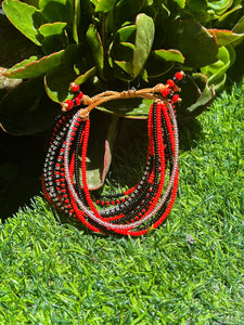 The Shanga Slide Beaded Bracelet by Her Best Foot Forward, inspired by Maasai jewelry, features vibrant red and black beads and is shown on green grass with leafy plants and sunlight highlighting its East African design.