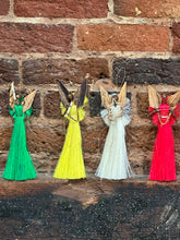 Load image into Gallery viewer, Four Angels From On High ornaments by Her Best Foot Forward—handcrafted fiber angels in green, yellow, white, and red dresses—stand against a rustic brick wall. Made in Tanzania by women artisans.