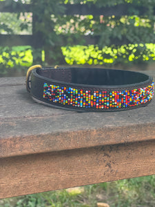 The Buddy Beaded Dog Collar by Her Best Foot Forward, a classic handmade Maasai pet accessory from East Africa, features colorful beadwork on dark leather and rests outdoors on weathered wood with blurred green foliage in the background.