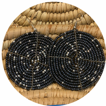Load image into Gallery viewer, Two large, round Duara Circular Beaded Earrings by Her Best Foot Forward, handmade Maasai statement jewelry from East Africa in black and silver beads, are displayed against woven straw, highlighting their striking spiral pattern.