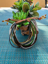 Load image into Gallery viewer, The Shanga Slide Beaded Bracelet by Her Best Foot Forward—a vibrant, handmade Maasai slip-on—hangs on driftwood adorned with succulents, displayed atop a woven blue placemat.