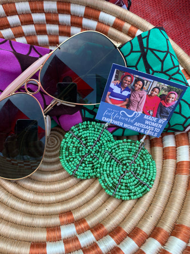 A pair of Her Best Foot Forward Duara Circular Beaded Earrings—handmade Maasai statement jewelry from East Africa—rests on a woven basket beside aviator sunglasses, colorful fabric, and a card reading “Made by Women Artisans to Empower Women & Girls.”.