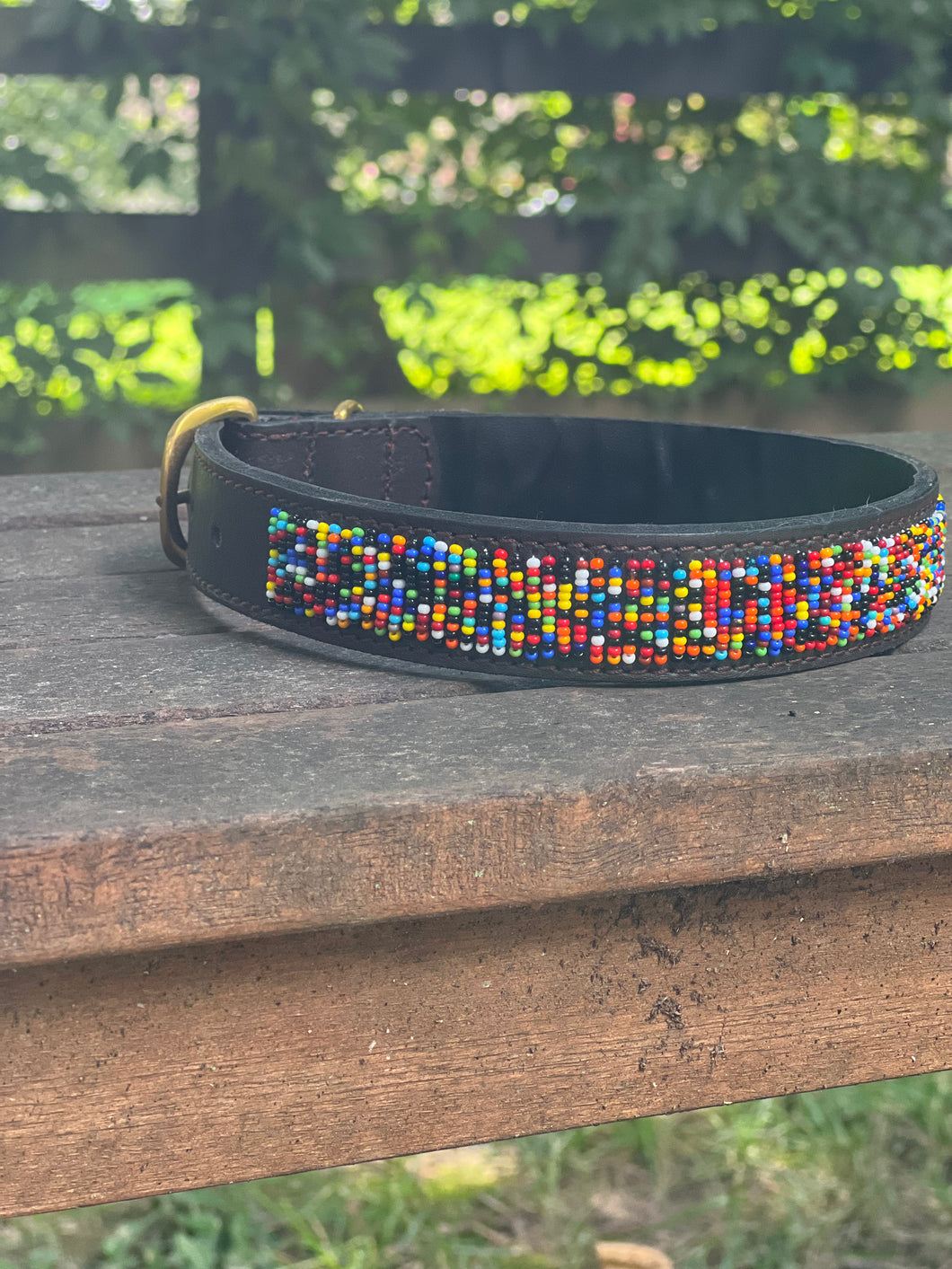 The Buddy Beaded Dog Collar by Her Best Foot Forward, a classic handmade Maasai pet accessory from East Africa, features colorful beadwork on dark leather and rests outdoors on weathered wood with blurred green foliage in the background.