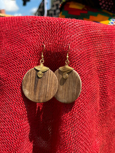 The Rosemiti Hand-Carved Rosewood Earrings by Her Best Foot Forward, accented with brass, are displayed against vibrant red fabric and colorful patterns, reflecting the rich heritage of East African statement jewelry.