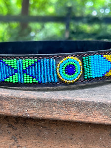A close-up of Her Best Foot Forward’s Proud as a Peacock Beaded Dog Collar, a handmade ethical Maasai pet accessory from East Africa, featuring vibrant blue, green, and yellow geometric beadwork on wood with blurred greenery behind.