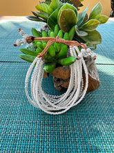 Load image into Gallery viewer, The Shanga Slide Beaded Bracelet by Her Best Foot Forward, an easy slip-on Maasai-inspired design from East Africa, features white beads and brown cord, displayed on a log amid green succulents atop a blue woven mat.