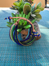 Load image into Gallery viewer, The Shanga Slide Beaded Bracelet by Her Best Foot Forward, with blue, green, and yellow strands inspired by Maasai tradition, is displayed on a succulent in a wooden holder atop turquoise weaving—celebrating East African artistry.
