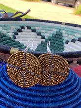 Load image into Gallery viewer, Her Best Foot Forward’s Duara Circular Beaded Earrings—handmade Maasai statement jewelry from East Africa—feature gold and dark blue designs, displayed before a woven basket with green, black, and white stripes in a blurred outdoor setting.