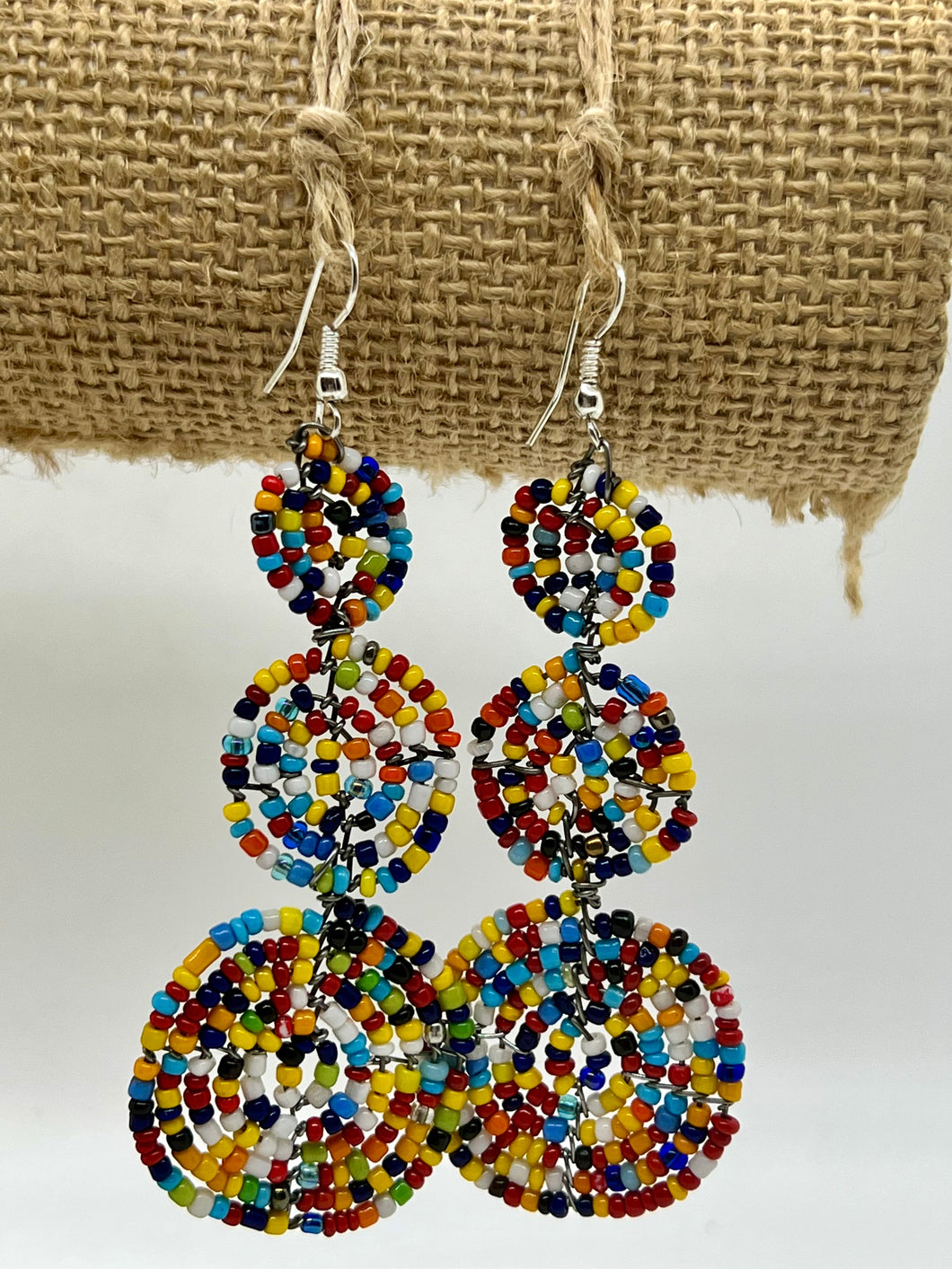 The Kiungo Conjoined Circle Earrings by Her Best Foot Forward are handmade Maasai statement jewelry from East Africa, featuring small, colorful beads in three linked circles, displayed against a textured burlap background.