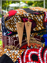 Load image into Gallery viewer, The Rosemiti Hand-Carved Rosewood Earrings from Her Best Foot Forward, featuring brass accents, hang on a vibrant patterned hat with greenery and blurred outdoor scenery in the background.