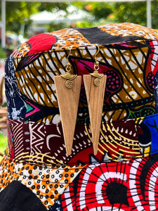 The Rosemiti Hand-Carved Rosewood Earrings from Her Best Foot Forward, featuring brass accents, hang on a vibrant patterned hat with greenery and blurred outdoor scenery in the background.