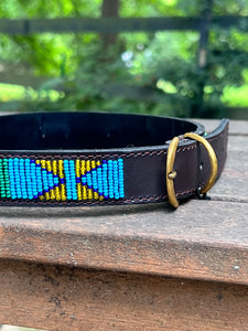 The Proud as a Peacock beaded dog collar by Her Best Foot Forward, handmade in East Africa with vibrant Maasai beadwork, is displayed on a wooden surface outdoors.