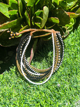 Load image into Gallery viewer, The Shanga Slide Beaded Bracelet by Her Best Foot Forward, inspired by Maasai jewelry from East Africa with black, white, and gold beads, is shown on green grass with leafy plants in bright sunlight.