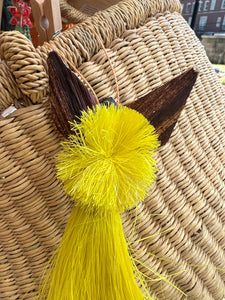 A close-up of an Angels From On High ornament by Her Best Foot Forward, handcrafted by Tanzanian women. The woven basket features two brown ears and a fluffy yellow pom-pom, with blurred buildings in the background.