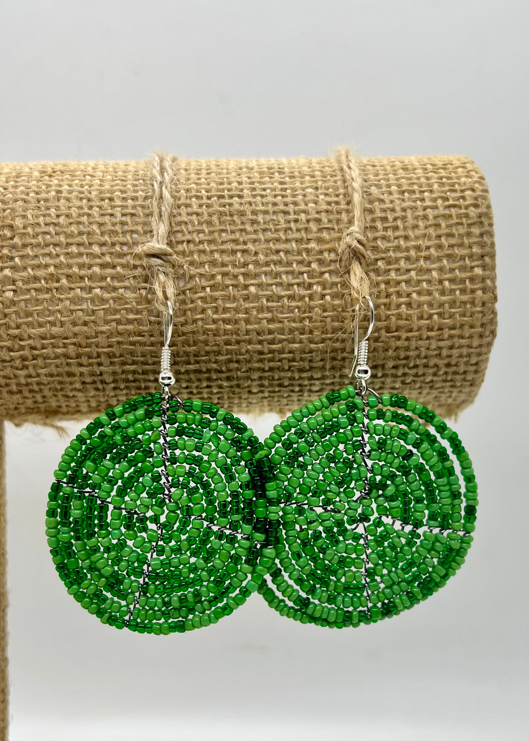 The Duara Circular Beaded Earrings by Her Best Foot Forward, handmade Maasai statement jewelry from East Africa, feature round green beads with peace sign designs and are displayed on twine against a white background.