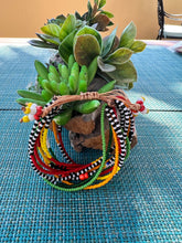 Load image into Gallery viewer, The Shanga Slide Beaded Bracelet by Her Best Foot Forward, inspired by Maasai jewelry with red, yellow, green, black, and white beads, is displayed in front of a small potted succulent on a blue woven placemat.
