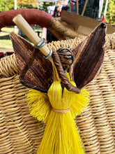 Load image into Gallery viewer, The Angels From On High ornament by Her Best Foot Forward is handmade in Tanzania from banana fiber, featuring a yellow raffia skirt, brown leaf wings, and a flute—beautifully displayed against a woven basket at an outdoor market.