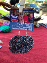 Load image into Gallery viewer, Duara Circular Beaded Earrings by Her Best Foot Forward, handmade Maasai statement jewelry in black and silver, hang on display with a sign promoting women artisans’ empowerment. A red cloth and outdoor market setting complete the scene.