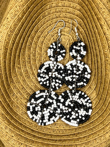 The Kiungo Conjoined Circle Earrings by Her Best Foot Forward are handmade Maasai statement earrings from East Africa, featuring black and white beads in spiral patterns, shown on a woven tan basket background.